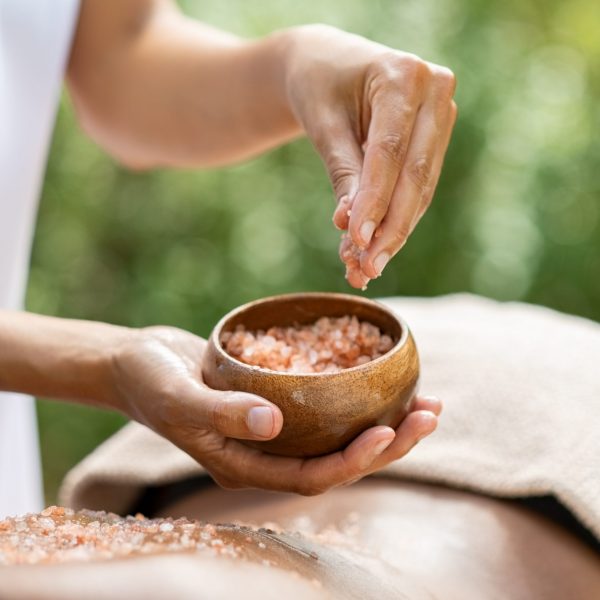 Closeup of woman lying and receiving massage with salt crystals at spa. Woman hand applying scrub on african woman back at wellness center for body exfoliation. Beauty therapist pouring salt for peeling on girl back.